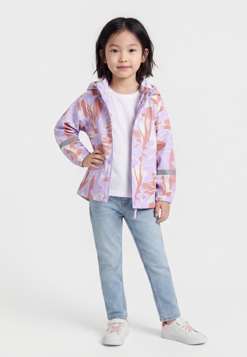 Νεαρή κοπέλα ντυμένη με παλ παστέλ ζακέτα με φλοράλ μοτίβα και φερμουάρ, λευκό t-shirt, ανοιχτό μπλε τζιν και λευκά αθλητικά παπούτσια με ροζ κορδόνια, στέκεται με το ένα χέρι στη μέση.