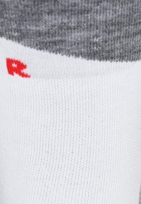 Acercamiento de un calcetín gris y blanco con una pequeña letra "R" roja tejida cerca del borde superior.