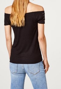 Czarny top typu off-the-shoulder z prążkowanej tkaniny, z krótkimi rękawami i detalami w postaci oczek. W zestawie z jasnoniebieskimi dżinsami z klasycznymi kieszeniami.