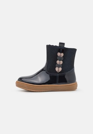 Bottine noire brillante avec semelle marron, dessus en tissu, et rangée décorative de formes de cœur alternant noir et beige sur le côté.