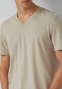 Beżowy t-shirt z dekoltem w serek, wykonany z gładkiego materiału, z krótkimi rękawami i subtelnym logo z przodu, dopasowany krój.