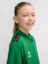 Młoda dziewczyna z jasnymi, związanymi do tyłu włosami, ubrana w zieloną sportową kurtkę Hummel, lekko się uśmiecha na jednolitym jasnym tle.