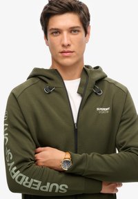 Felpa con zip verde oliva, dotata di cappuccio, zip nera e logo sul petto sinistro e sulla manica. Presenta dettagli con cordini e una texture morbida.