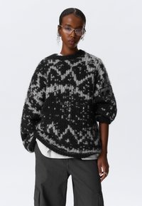 Pull oversized noir et gris à motif avec une texture douce, présentant un design géométrique et des manches larges, associé à un pantalon cargo noir.
