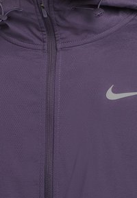 Lila perforierte Jacke mit frontalem Reißverschluss, verstellbarem Kapuzenzug und einem weißen Nike-Logo auf der rechten Brust.