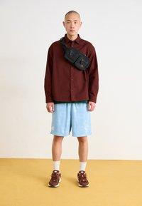 Burgundy gombos ing, zöld ing alatta, világoskék mintás rövidnadrág, fekete keresztpántos táska, barna tornacipő, fehér zokni. Egyszerű háttér.