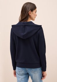 Haut à capuche bleu marine en tissu doux, avec une capuche spacieuse, des poignets et un ourlet côtelés. La vue arrière montre un design simple et épuré.