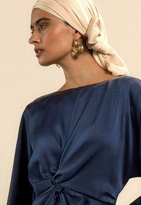 Tumesinine pluus, millel on vöökoha juures keeruline sõlm, valmistatud siledast kangast. Modellil on õlakreem värvi pearätt ja kuldsed rõngaskõrvarõngad.