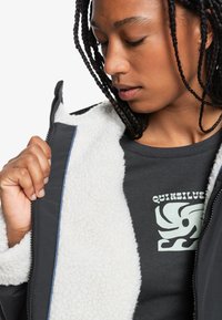 Čierna bunda s bielou, huňatou podšívkou; obsahuje zips. Pod ňou je šedá košeľa, na ktorej je svetlozelený grafický potlač.