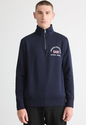 Marineblå hettegenser med høyt hals og halv glidelås. Har hvitt og rødt brodert logo på brystet. Glatt tekstur, ribbede mansjetter.