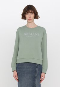 Světle zelená mikina vyrobená z měkké tkaniny, s kulatým výstřihem a žebrovanými manžetami. Na přední straně se nachází lesklé logo "ARMANI EXCHANGE".