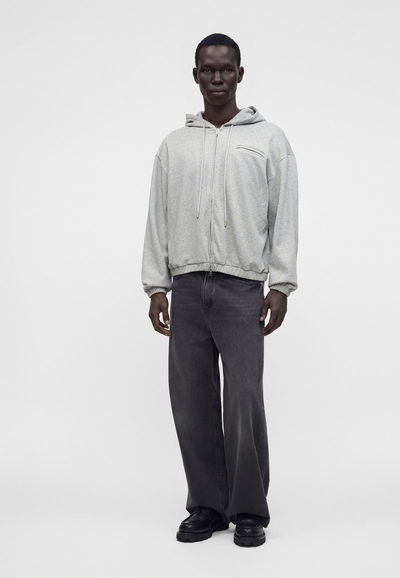Homme portant un sweat à capuche gris clair zippé, un jean gris foncé ample et des chaussures noires, debout devant un fond blanc uni.