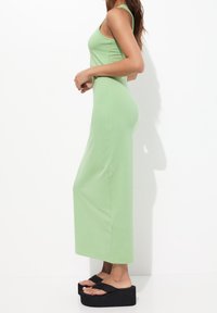 Kobieta stoi bokiem w bezrękawnej, jasnoszarej sukience maxi z czarnymi sandałami na platformie, na tle białym.