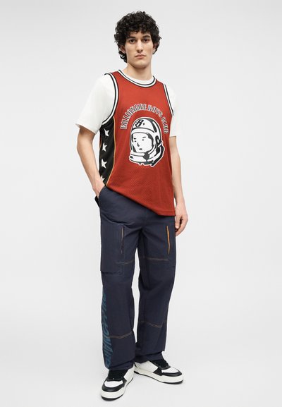 Νέος άνδρας που φοράει κόκκινο αμάνικο μπασκετικό φανελάκι με γραφικό αστροναύτη, λευκή μπλούζα, μπλε παντελόνι cargo και μαύρα-άσπρα αθλητικά παπούτσια.