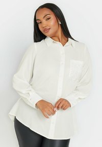 Camicia bianca a maniche lunghe con colletto button-down, orlo curvo e tasca decorata con piccole strass. Tessuto dalla consistenza liscia.