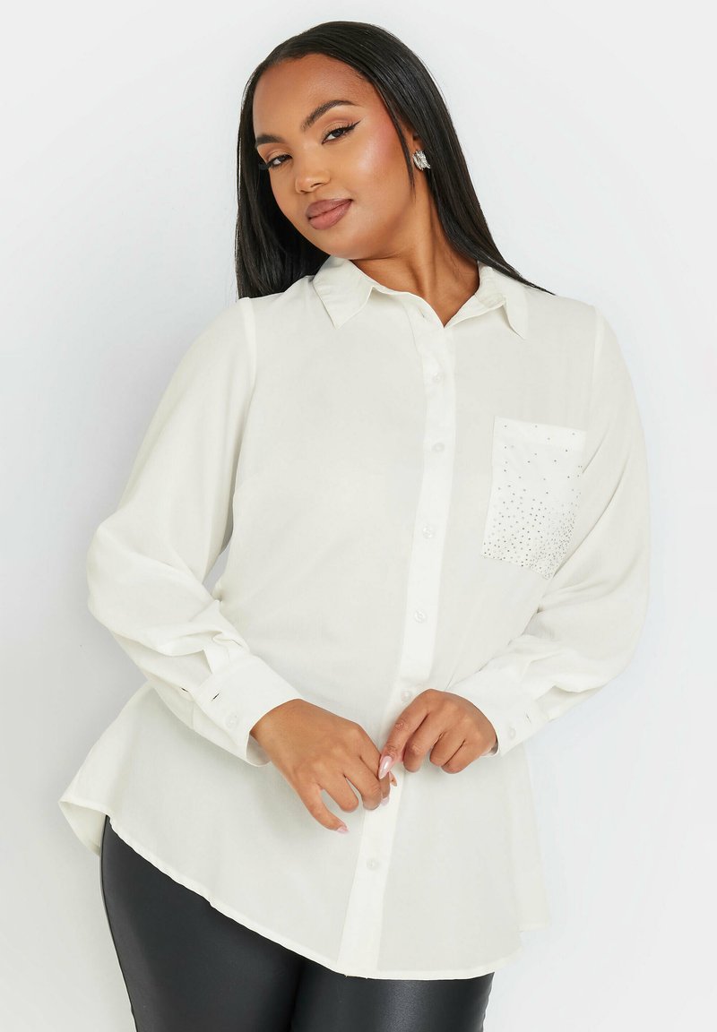 Camicia bianca a maniche lunghe con colletto button-down, orlo curvo e tasca decorata con piccole strass. Tessuto dalla consistenza liscia.