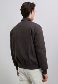 Hombre vistiendo una chaqueta bomber marrón oscuro con puños y cinturilla acanalados, combinada con pantalones de color claro, de pie contra un fondo plano.