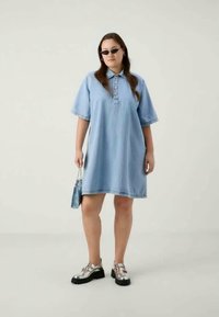 Ανοιχτό μπλε τζιν φόρεμα-πουκάμισο με κουμπωτή πατιλέτα, κοντά μανίκια και χαλαρή εφαρμογή. Φοριέται με ασημένια παπούτσια πλατφόρμας και μικρή τσάντα.