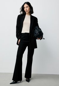 Black blazer over a light beige ribbed top, paired with black tailored pants and pointed-toe heels. A black drawstring bag is included.