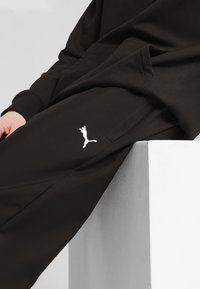 Juodos medvilnės treningo kelnes su baltu Puma logo ant šlaunies, šoninės kišenės ir laisvas prigludimas.