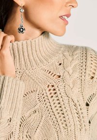 Frau trägt einen beigefarbenen Kabelstrickpullover und silberne Ohrhänger in Rosenform, Hand berührt ihren Hals nahe dem Kragen.