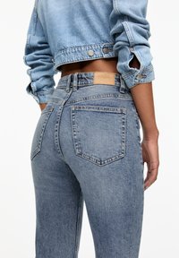 Jeans de mezclilla azul claro con una cintura media, que cuentan con dos bolsillos traseros, un parche de cuero marrón y una textura desgastada.