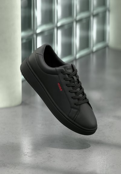 Baskets basses noires avec lacets noirs et logo rouge "HUGO" sur le côté, suspendues au-dessus d'un sol gris réfléchissant.