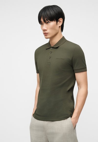 Νεαρός άνδρας με κοντομάνικο πουκάμισο πόλο σε ελιά πράσινο και ανοιχτό γκρι καρό παντελόνι, στέκεται με τα χέρια στις τσέπες, ουδέτερο φόντο.