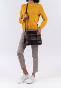 Kobieta w musztardowym swetrze, w kratkowanych spodniach, w białych trampkach, niosąca czarną torebkę na ramię o fakturowanej powierzchni z frędzlem.
