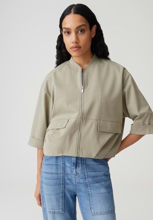 Frau mit langen, dunklen lockigen Haaren, die eine beigefarbene Jacke mit Reißverschluss, Klappentaschen und umgeschlagenen Ärmeln trägt, kombiniert mit hoch taillierten blauen Jeans.