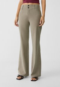 Vrouw in een beige en zwarte geruite flared broek met een hoge taille en zwarte sandalen met hakken en dunne bandjes.