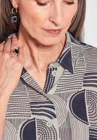 Hemdbluse in Hellgrau mit marineblauen geometrischen Mustern, ausgestattet mit einem Kragen, Knöpfen und glatter Textur; begleitet von kreisförmigem Schmuck.