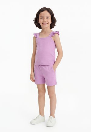 Dievča stojí a usmieva sa, má na sebe lilavý tielko bez rukávov s volánmi na ramenách, zodpovedajúce krátke nohavice a biele tenisky na bielom pozadí.