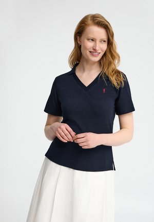 Kobieta z rudymi włosami, ubrana w granatową koszulkę polo z krótkim rękawem i białą plisowaną spódnicę, uśmiechająca się i delikatnie patrząca na bok.