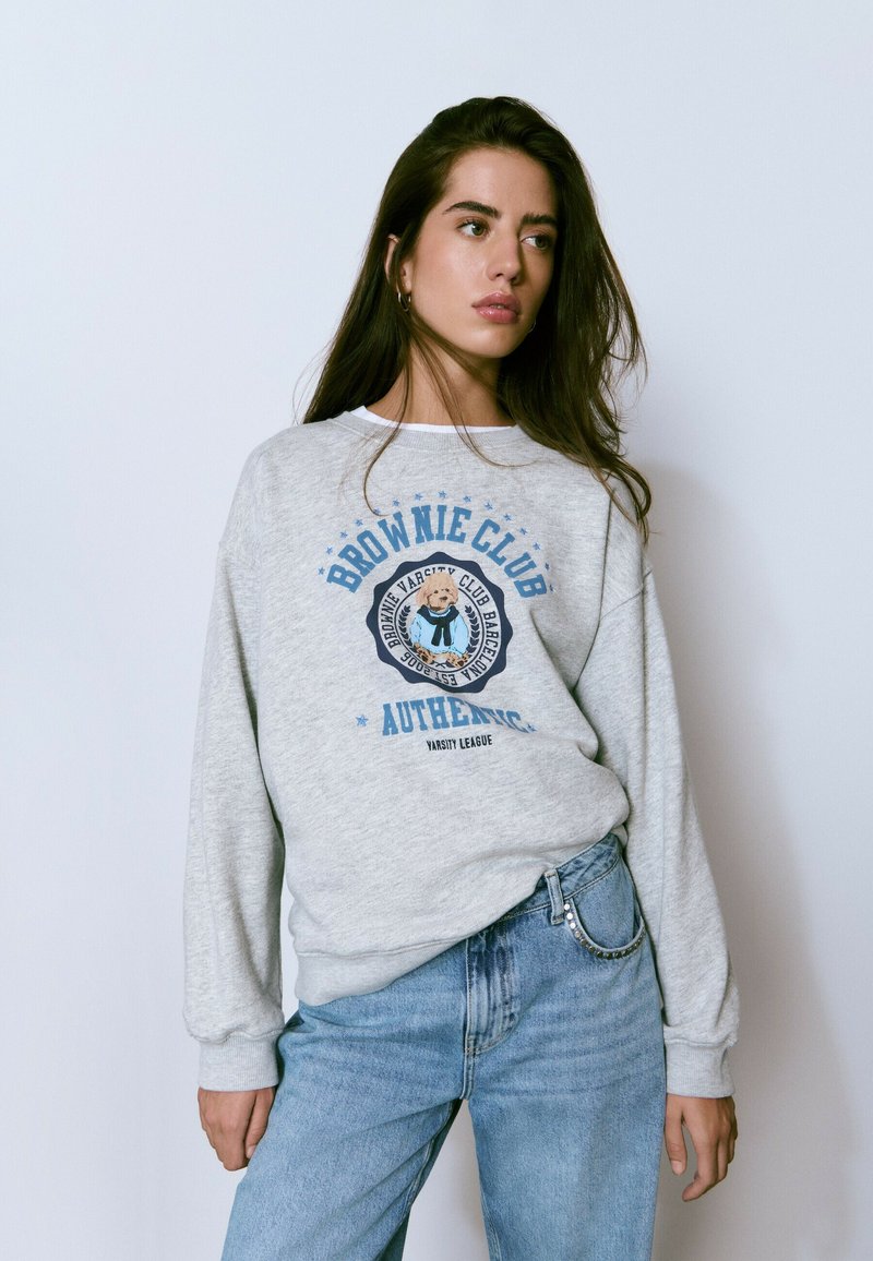 Joven con cabello largo y castaño, vistiendo un sudadera gris con un gráfico de perro y jeans azules con detalles de tachuelas en el bolsillo.