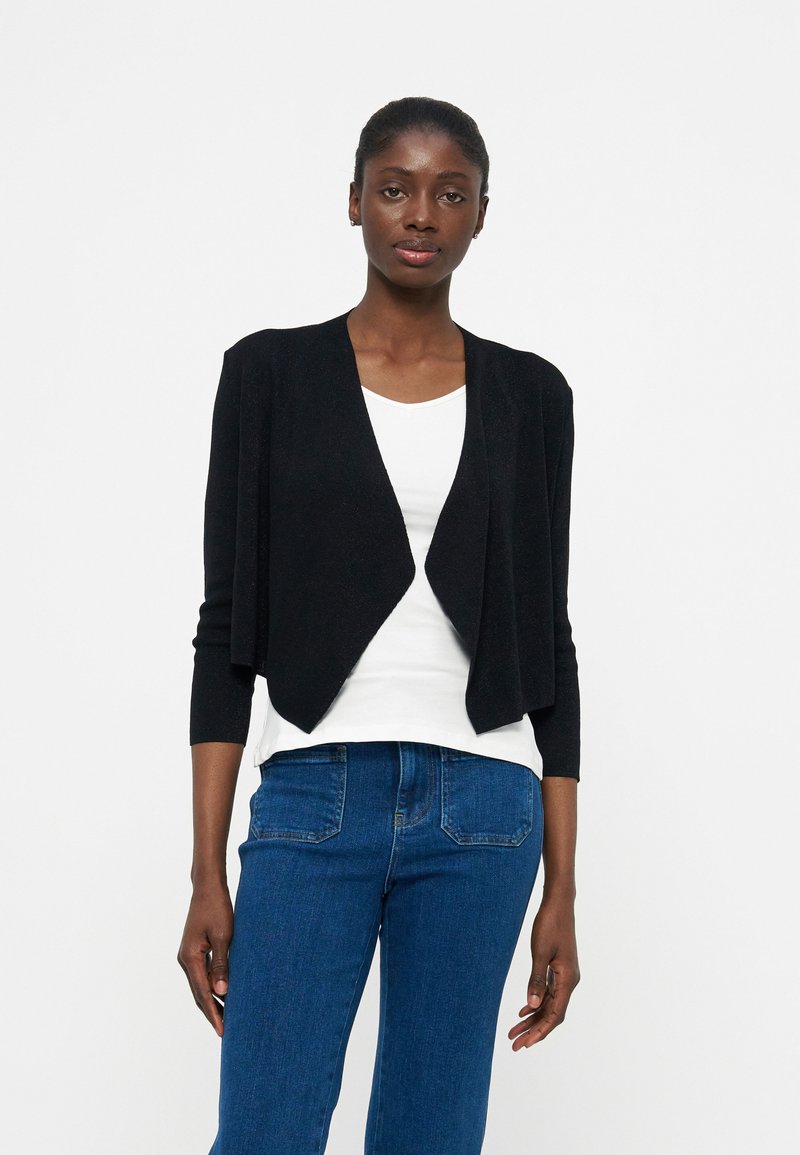 Γυναίκα που φοράει μαύρη ζακέτα με ανοικτό μπροστινό μέρος πάνω από λευκή μπλούζα και μπλε τζιν, στέκεται μπροστά σε απλό λευκό φόντο.