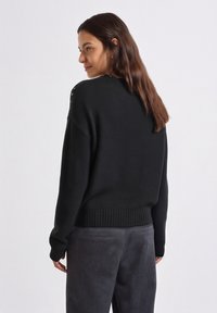 Zwarte gebreide trui met geribbelde zoom en manchetten; voorzien van subtiele details op de schouders, gecombineerd met grijze getextureerde broek.