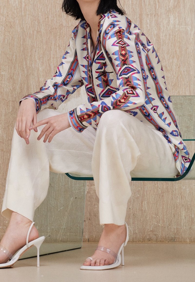 Chemise à manches longues avec un motif géométrique multicolore en rouge, bleu et crème ; associée à un pantalon large blanc texturé et des sandales à talons transparents.