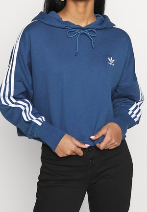 Personne portant un sweat à capuche Adidas bleu avec des bandes blanches sur les manches, un détail à nouer au niveau du cou et un pantalon noir avec fermeture par boutons visible.