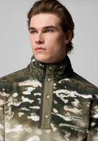 Pullover in pile verde e beige con colletto alto, bottoni a pressione e tasca con zip. Presenta un motivo camouflage e una texture morbida.