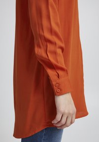 Langärmlige Bluse aus lebhaftem orangefarbenem Stoff, mit einem geknöpften Ärmelabschluss, der zwei Knöpfe hat, und einem leicht geschwungenen Saum f�ür einen entspannten Stil.