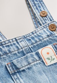 Salopette en jean bleu clair avec des boutons marron et un petit patch blanc avec une fleur de marguerite sur la poche avant.