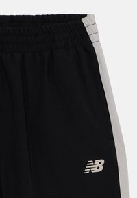Pantalones deportivos negros con cinturilla elástica, franja lateral gris y un pequeño logo blanco en la parte baja de la pierna. Textura de tela suave.