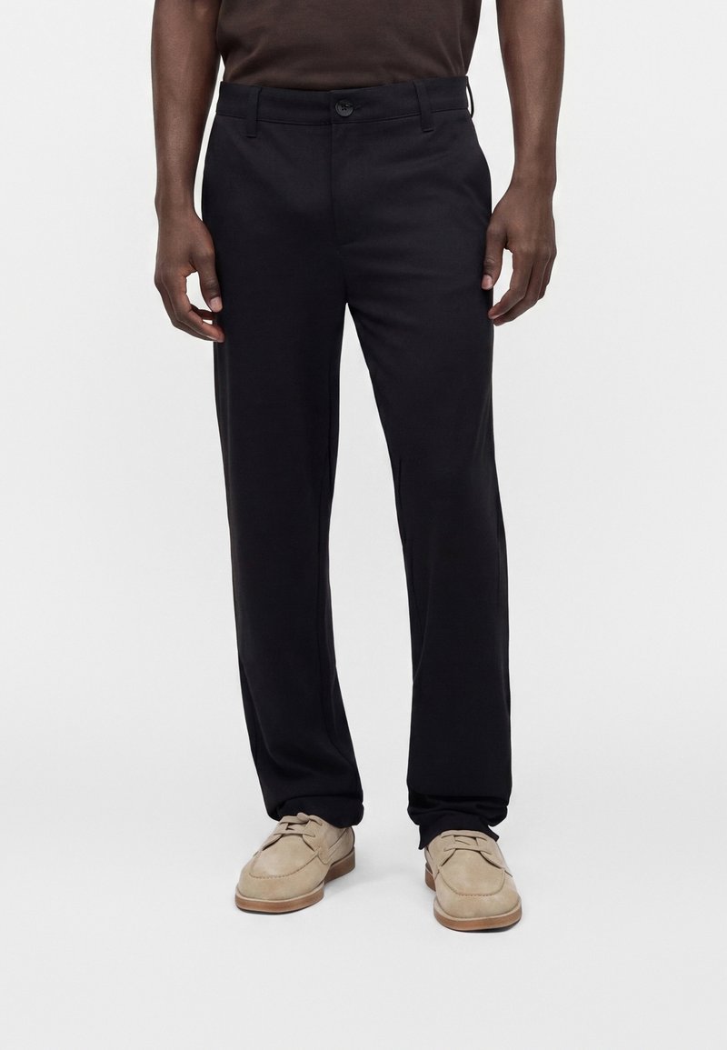 Homme portant un pantalon noir droit et des chaussures beiges en daim à lacets, debout sur un fond blanc.