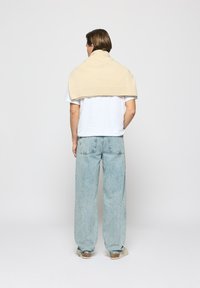 Beige gestrickter Pullover über den Schultern einer Person, die ein weißes T-Shirt und eine helle, weit geschnittene Jeans trägt.