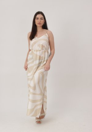 Kobieta o długich ciemnych włosach, ubrana w bezrękawnikową, beżowo-białą suknię maxi w wzory i sandały na obcasie z odkrytymi palcami, stoi na tle jednolitego tła.