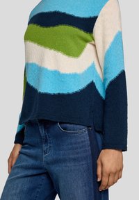 Person, der einen mehrfarbigen gestreiften Pullover in Blau, Grün, Creme und Marineblau sowie dunkelblaue Jeans trägt, vor einem einfarbigen Hintergrund.