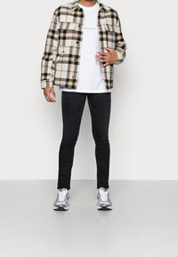 Camisa de franela a cuadros en crema, negro y naranja, usada sobre una camiseta blanca, combinada con jeans ajustados negros y zapatillas grises.