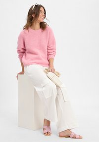 Vrouw zittend op een blok, gekleed in een roze trui, witte wijde broek, roze sandalen, met een kleine beige handtas, kijkt opzij en glimlacht.