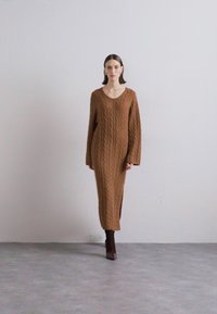 Hnedé dlhé šaty s pleteným vzorom a V výstrihom, dlhými rukávmi, bočnými rozparkami a voľným strihom, skombinované s hnedými podpätkovými čižmami.